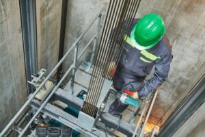 A lift machinist repairing an elevator inside a lift shaft, surrounded by mechanical components and tools.