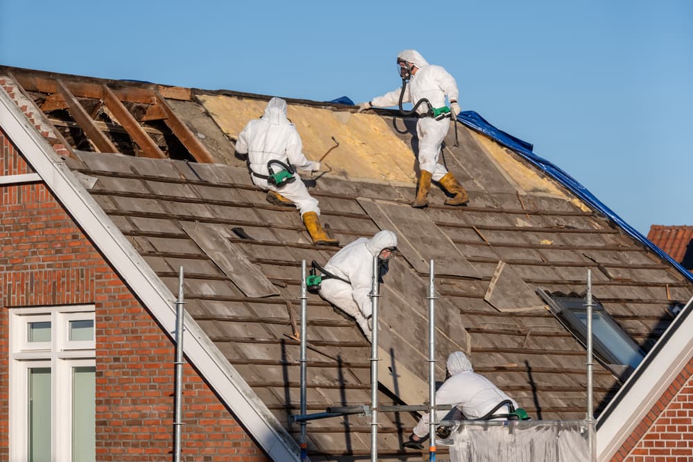Professionals in protective suits are removing asbestos-cement roofing underlayment.