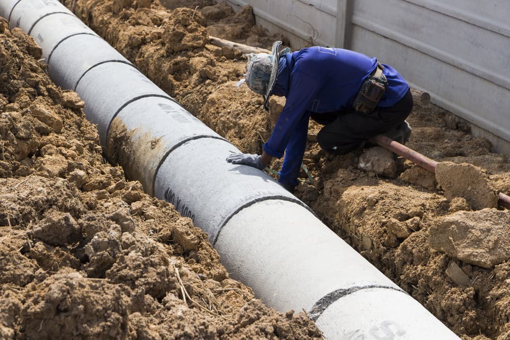 A worker installs precast concrete manholes for storm water drainage, working outdoors in hot weather against a construction industry backdrop.