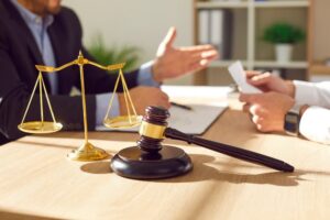 A close-up photo of a male lawyer at his office desk, engaged in discussion with a male client. The desk features a judge's gavel and scales, symbolizing justice. 