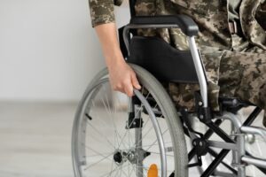 A military woman is seated in a self-propelled manual wheelchair indoors. She is dressed in camouflage attire, but her face is unrecognizable, emphasizing her role as a soldier.